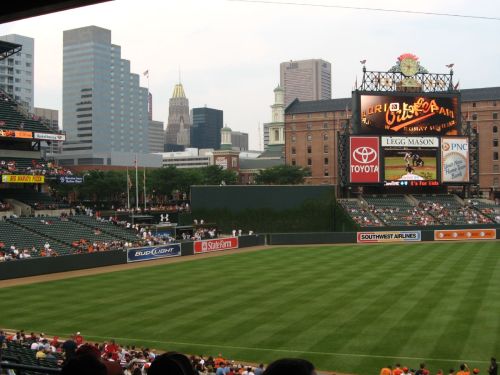 A baseball stadium with a green field, city skyline in the background, and a large scoreboard displaying advertisements.