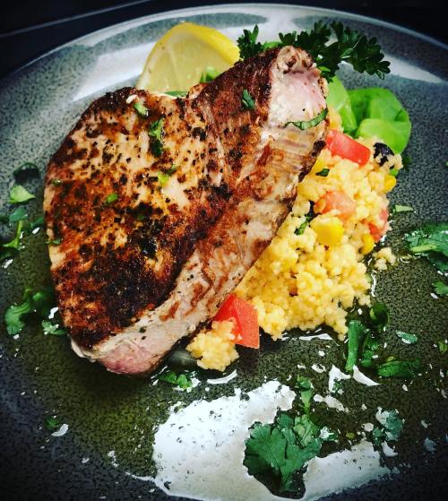 Grilled fish fillet on a plate with couscous, vegetables, and a lemon wedge, garnished with herbs.