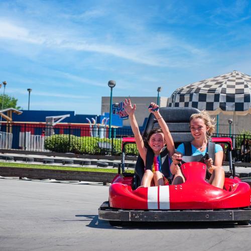 Two girls joyfully ride in a red go-kart, raising their hands in excitement on a sunny day at an amusement park.