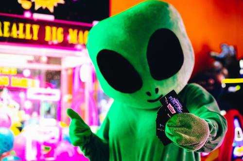 A green alien mascot holds game tickets, smiling in a colorful arcade setting.