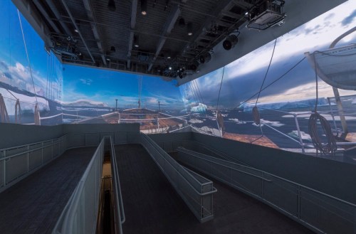 A spacious room with large screens displaying scenic landscapes and a ramp leading downwards.