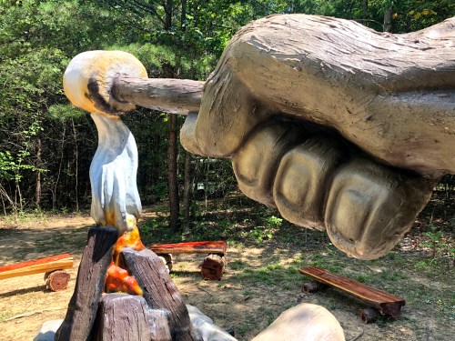 A large hand sculpture striking a log over a campfire, surrounded by trees and wooden benches.