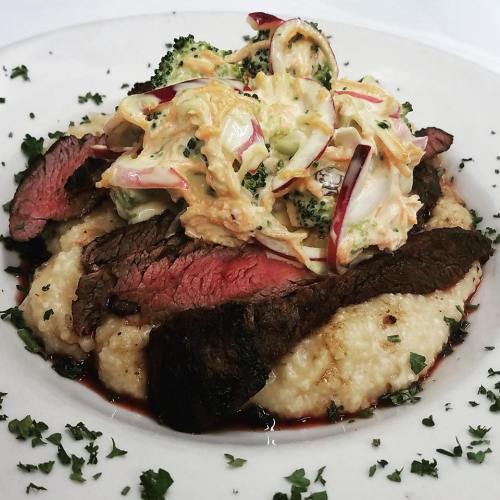 A plate of creamy grits topped with sliced beef and a colorful broccoli salad, garnished with parsley.