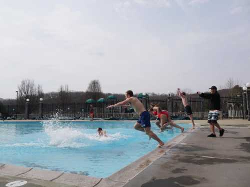 A group of boys jumps into a pool on a sunny day, creating splashes as they dive in.