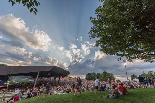 A large crowd gathers at an outdoor concert venue under a dramatic sky with rays of sunlight breaking through the clouds.