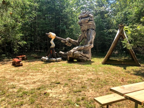 A large wooden sculpture of a giant figure roasting marshmallows over a campfire in a forested area.