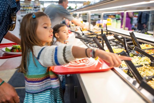 Two children at a buffet, one pointing at food options while holding a red tray, with adults in the background.