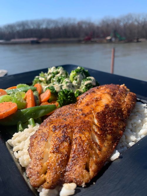 A plate of seasoned fish fillet on rice, accompanied by steamed broccoli and mixed vegetables, with a river view in the background.