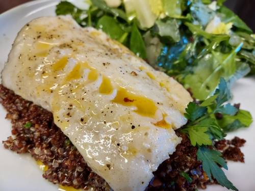 A plate featuring a piece of fish drizzled with olive oil, served on quinoa with a side of fresh salad.