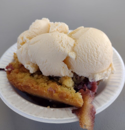 A dessert featuring a warm cookie topped with three scoops of vanilla ice cream on a paper plate.