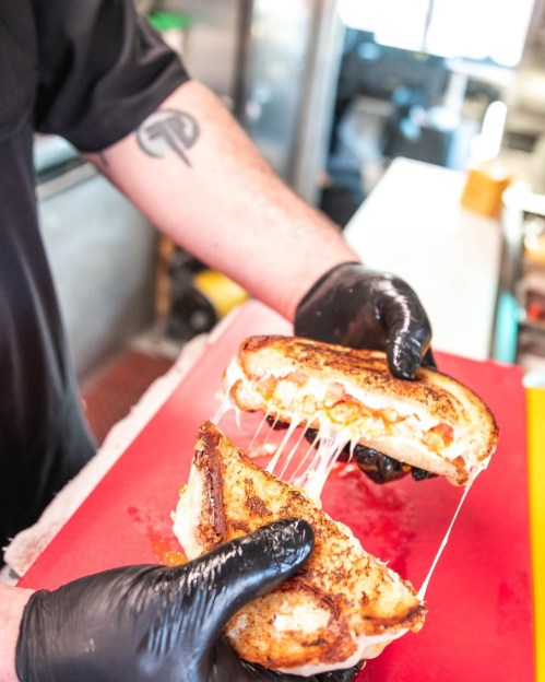 A chef in black gloves pulls apart a grilled cheese sandwich, revealing gooey melted cheese inside.