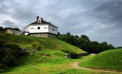 A historic white building on a grassy hill, surrounded by trees under a cloudy sky. A path leads towards the structure.