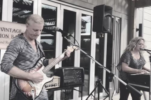 A man plays guitar while a woman sings into a microphone at an outdoor music venue.