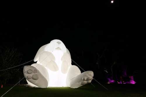 A large, illuminated inflatable rabbit sculpture sits in a dark outdoor setting, with a moon visible in the background.