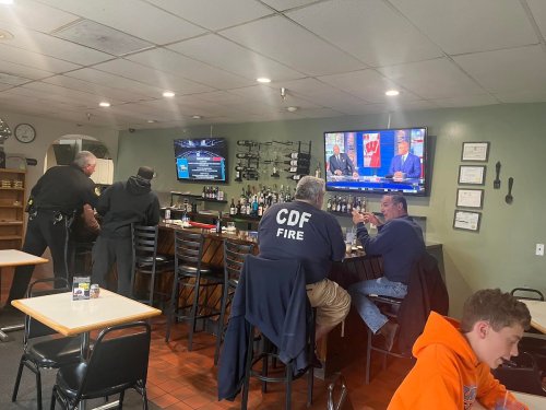 A bar scene with patrons seated at the counter, two TVs showing sports, and staff serving drinks.