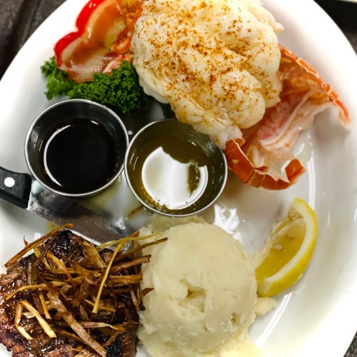 A plate featuring lobster tail, steak, mashed potatoes, lemon, and dipping sauces, garnished with greens.