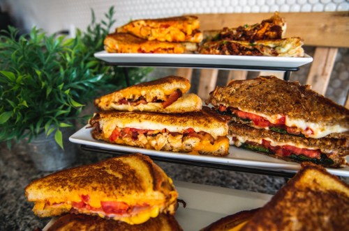 A display of grilled cheese sandwiches with various fillings, served on a tiered stand, alongside a small green plant.