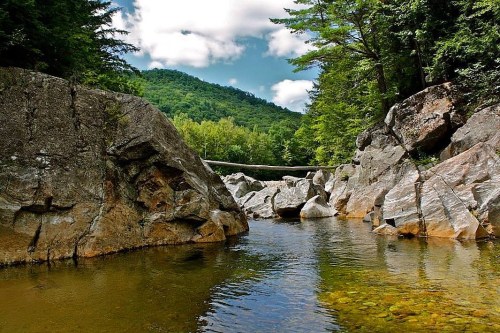 A serene river flows between large rocks, surrounded by lush green trees and a mountain under a partly cloudy sky.