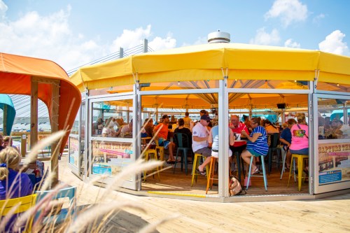A vibrant outdoor dining area with people enjoying meals under a yellow tent on a sunny day.