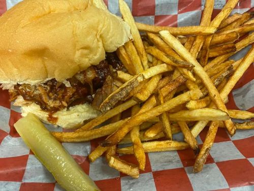 A pulled pork sandwich with barbecue sauce, served with a side of golden fries and a pickle on a red-checkered tray.