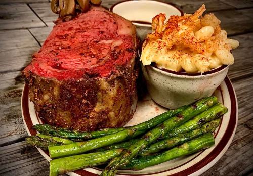 A plate featuring a juicy roast beef slice, asparagus, macaroni and cheese, and a small bowl of sauce.