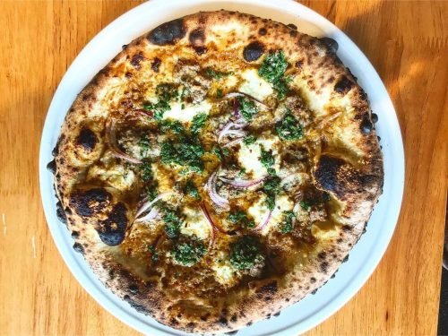 A freshly baked pizza topped with cheese, herbs, red onions, and a savory meat topping on a wooden table.