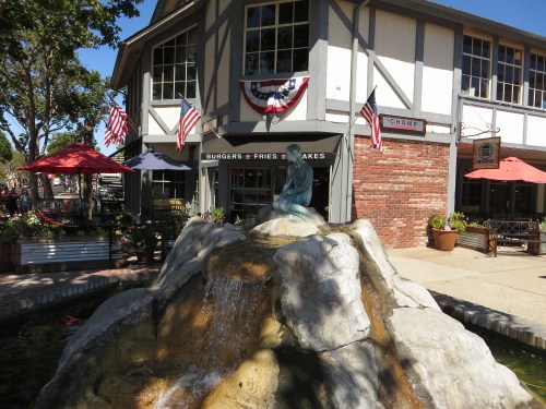 A fountain with a statue sits in front of a restaurant adorned with American flags and outdoor seating.