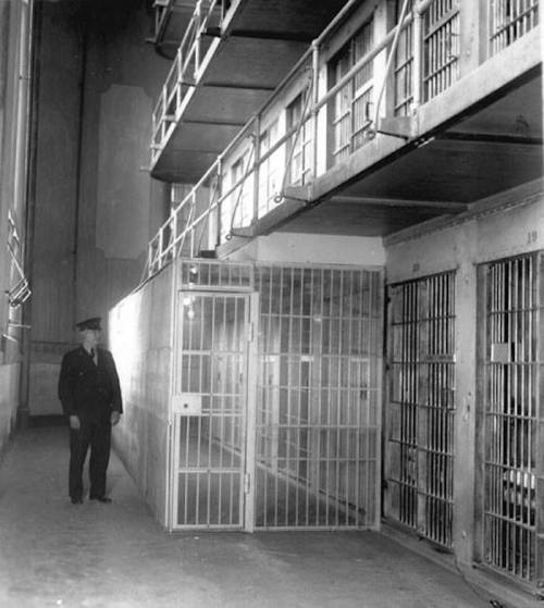 A black and white photo of a prison interior, featuring barred cells and a guard standing near a cell door.
