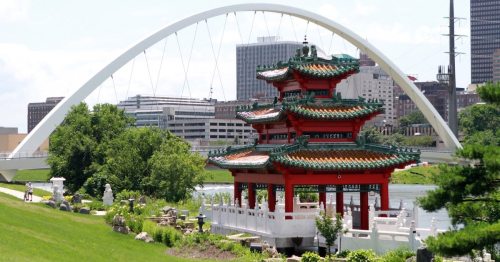 A traditional red pagoda surrounded by greenery, with a modern arch bridge and city skyline in the background.