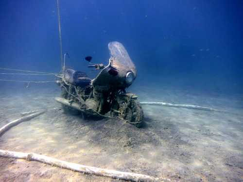 An old motorcycle submerged underwater, covered in mud and surrounded by wooden debris.