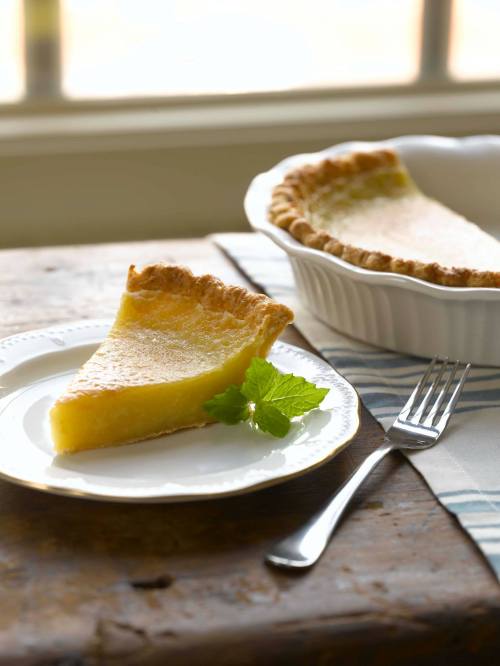 A slice of lemon pie on a plate, garnished with mint, next to a pie dish on a wooden table.