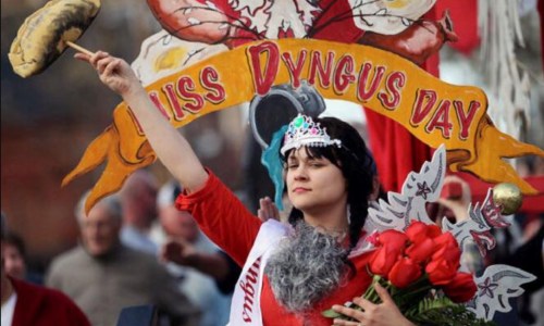 A woman in a tiara and red dress holds a potato and flowers, celebrating Dyngus Day with a festive banner in the background.