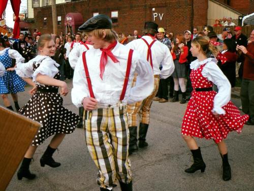 A lively dance scene with people in colorful polka dot outfits and suspenders, performing in a festive outdoor setting.
