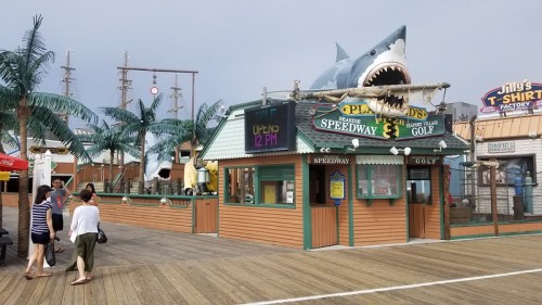A boardwalk scene featuring a mini-golf attraction with a large shark and palm trees, bustling with visitors.