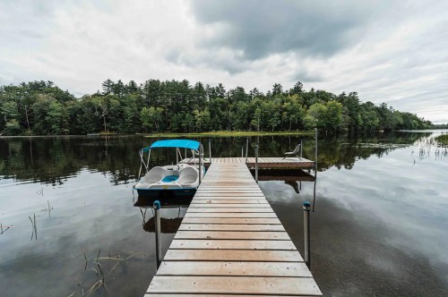 A wooden dock extends over a calm lake, with a blue boat tied nearby and lush green trees in the background under cloudy skies.