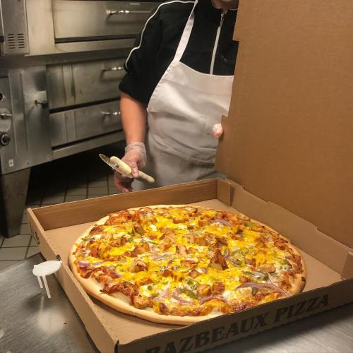 A freshly baked pizza with toppings in a takeout box, with a person preparing it in a kitchen.