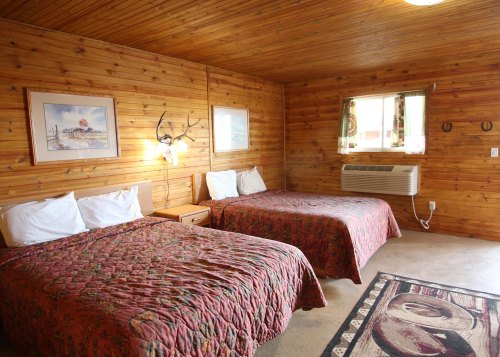 Cozy wooden room with two beds, a window, and rustic decor, featuring a patterned rug and wall art.