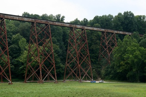 A tall, rusty railway trestle bridge stands over a green field, surrounded by dense trees under a cloudy sky.
