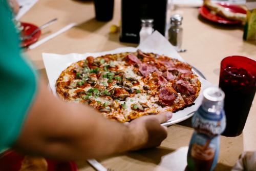 A person holds a large pizza topped with pepperoni, green peppers, and mushrooms, surrounded by drinks and plates.