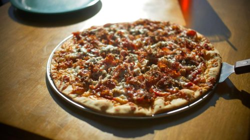 A freshly baked pizza topped with cheese, meat, and tomato sauce on a wooden table, with sunlight shining on it.