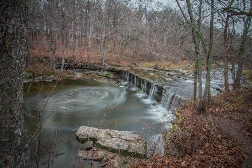 A serene waterfall cascades over rocks, surrounded by bare trees and autumn foliage in a tranquil forest setting.