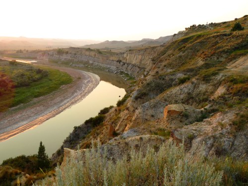 A winding river flows through a rugged landscape at sunset, with cliffs and greenery lining the banks.