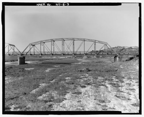 Black and white image of a metal arch bridge over a dry riverbed, surrounded by sparse vegetation and hills in the background.