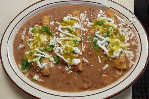 A plate of three enchiladas topped with green sauce, sour cream, cheese, and cilantro, served with refried beans.