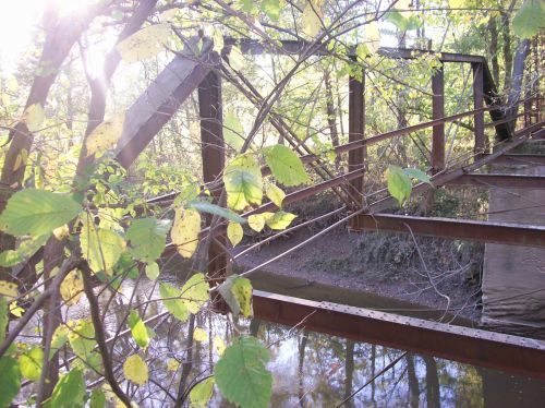 Rusty bridge structure partially obscured by green leaves, with a calm river reflecting sunlight in the background.