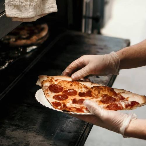 A hand in gloves holds a slice of pepperoni pizza on a paper plate, near an oven.