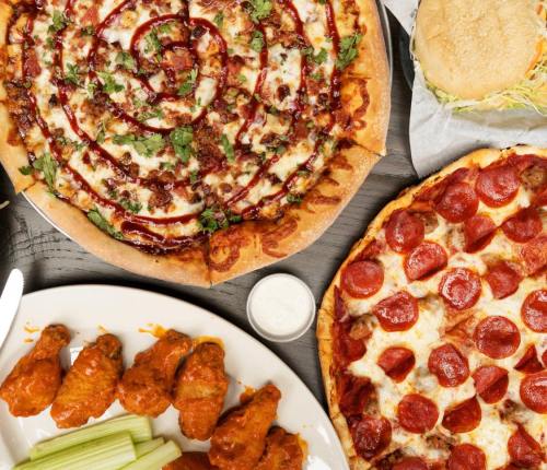 A spread of food featuring a BBQ chicken pizza, pepperoni pizza, buffalo wings, celery, and a burger.