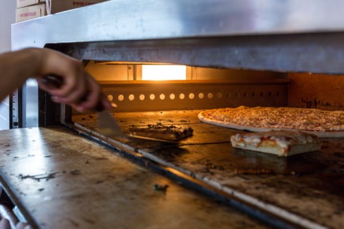 A hand using a spatula to remove pizza from a hot oven, with another pizza baking inside.
