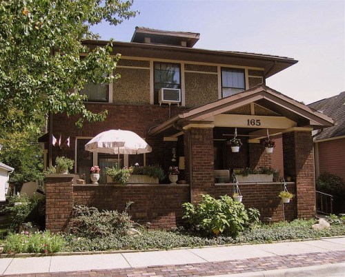 A charming two-story brick house with a porch, flower pots, and an umbrella, surrounded by greenery.
