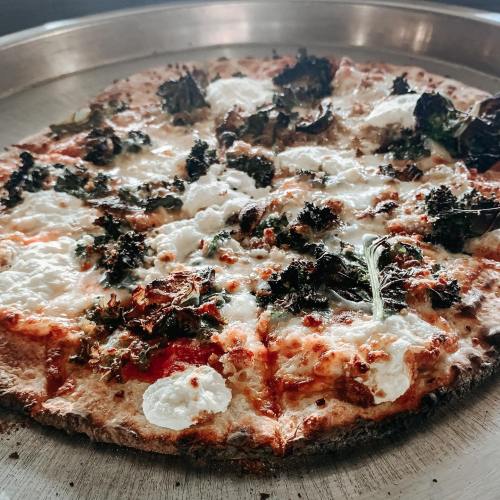 A close-up of a freshly baked pizza topped with cheese, kale, and dollops of ricotta on a metal pizza pan.